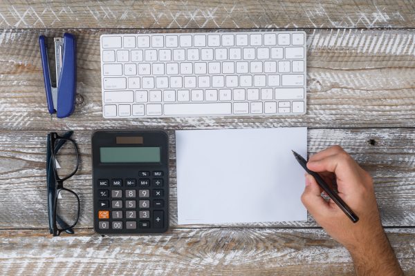 Top view man writing something on paper, with keyboard, eyeglasses, pen, cactus, magnifier on wooden background. horizontal